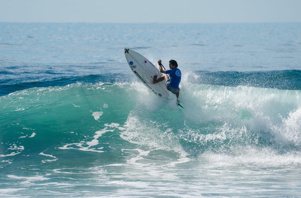 SUP Surfing | 奄美大島サーフィンスクールと体験、 サップ体験とシュノーケルツアーKazbo Surfer in a blue shirt rides a turquoise ocean wave, spraying water as the board cuts through the lip.